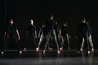 A group of dancers with their backs facing the camera and wearing black with a black background