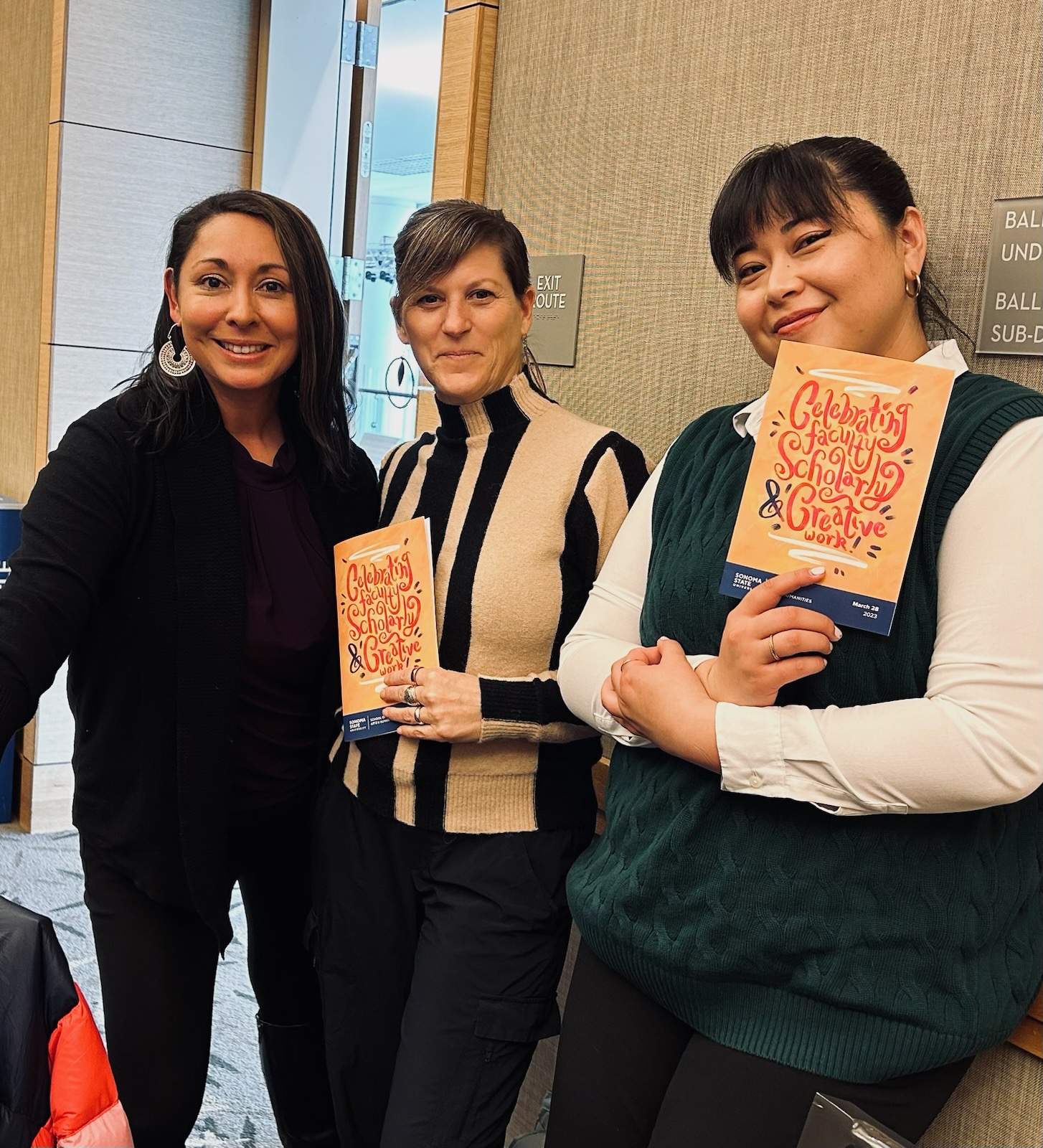 Marie Ramirez Downing, Christine Cali, and Farrah McAdam holding programs for "Celebrating Faculty Scholarly Creative Work!" event