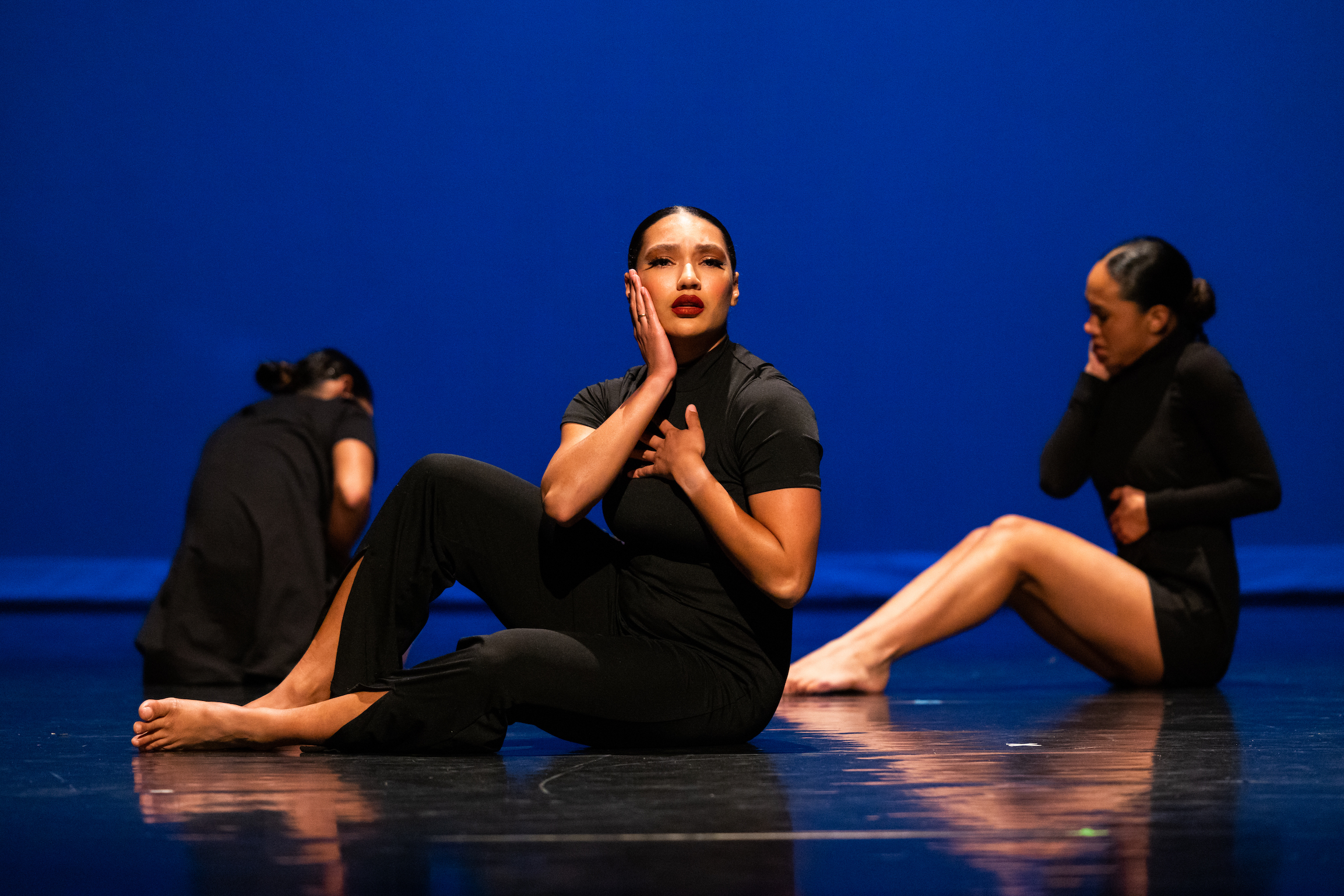 three dancers in black sitting on stage with a blue screen behind them