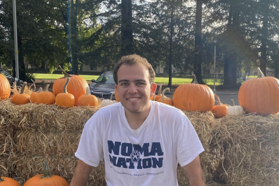 Avery Flasher-Duzgunes sitting on haystacks with some pumpkins around and in front of some trees wearing a 'Noma Nation' shirt