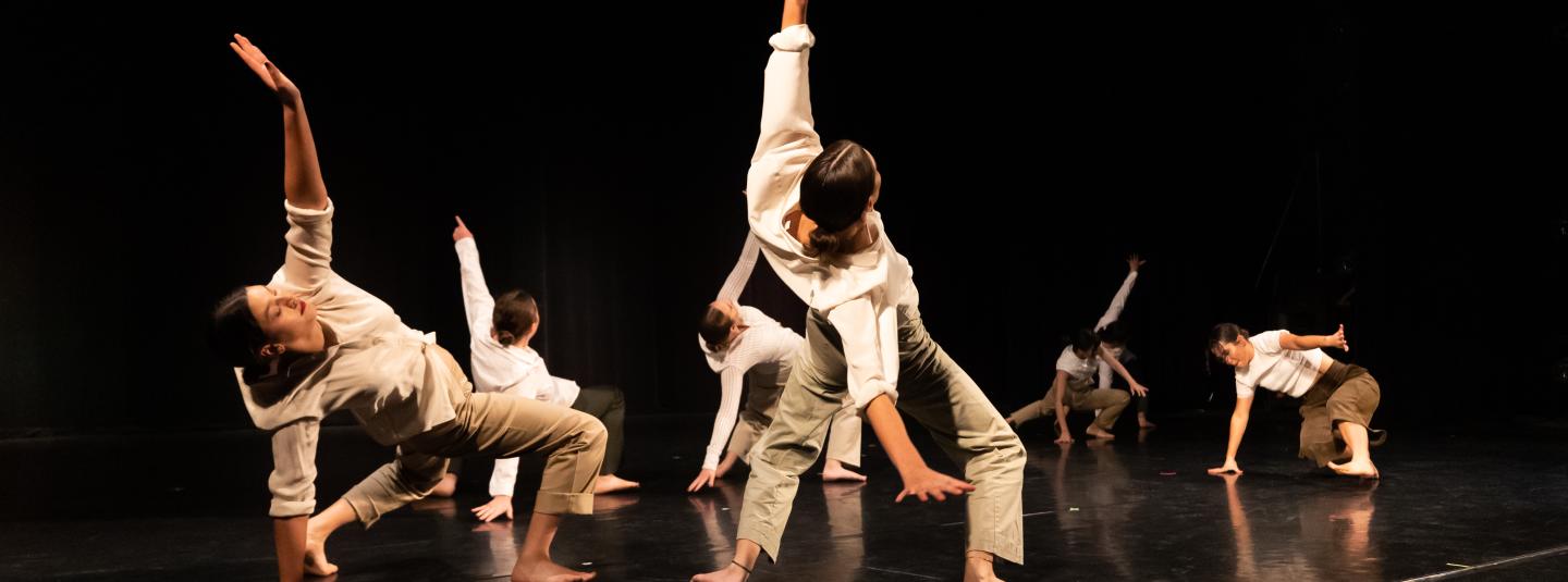 dancers in white shirts and beige pants leaning back on a dark stage