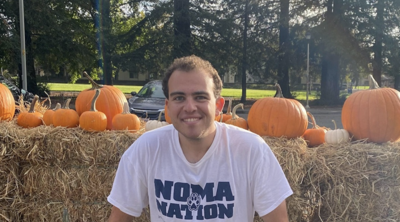 Avery Flasher-Duzgunes sitting on haystacks with some pumpkins around and in front of some trees wearing a 'Noma Nation' shirt