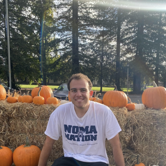 Avery Flasher-Duzgunes sitting on haystacks with some pumpkins around and in front of some trees wearing a 'Noma Nation' shirt